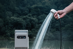 Hand holding white shower head spraying water outdoors, grey portable gas water heater on ground, green forest background, no text