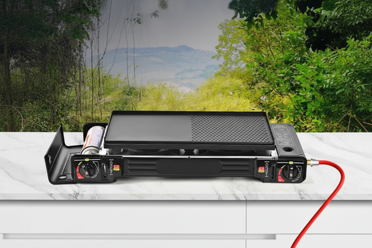 Black portable double burner camping gas stove on white marble surface with forest backdrop, red hose to propane cylinder, no text or watermark