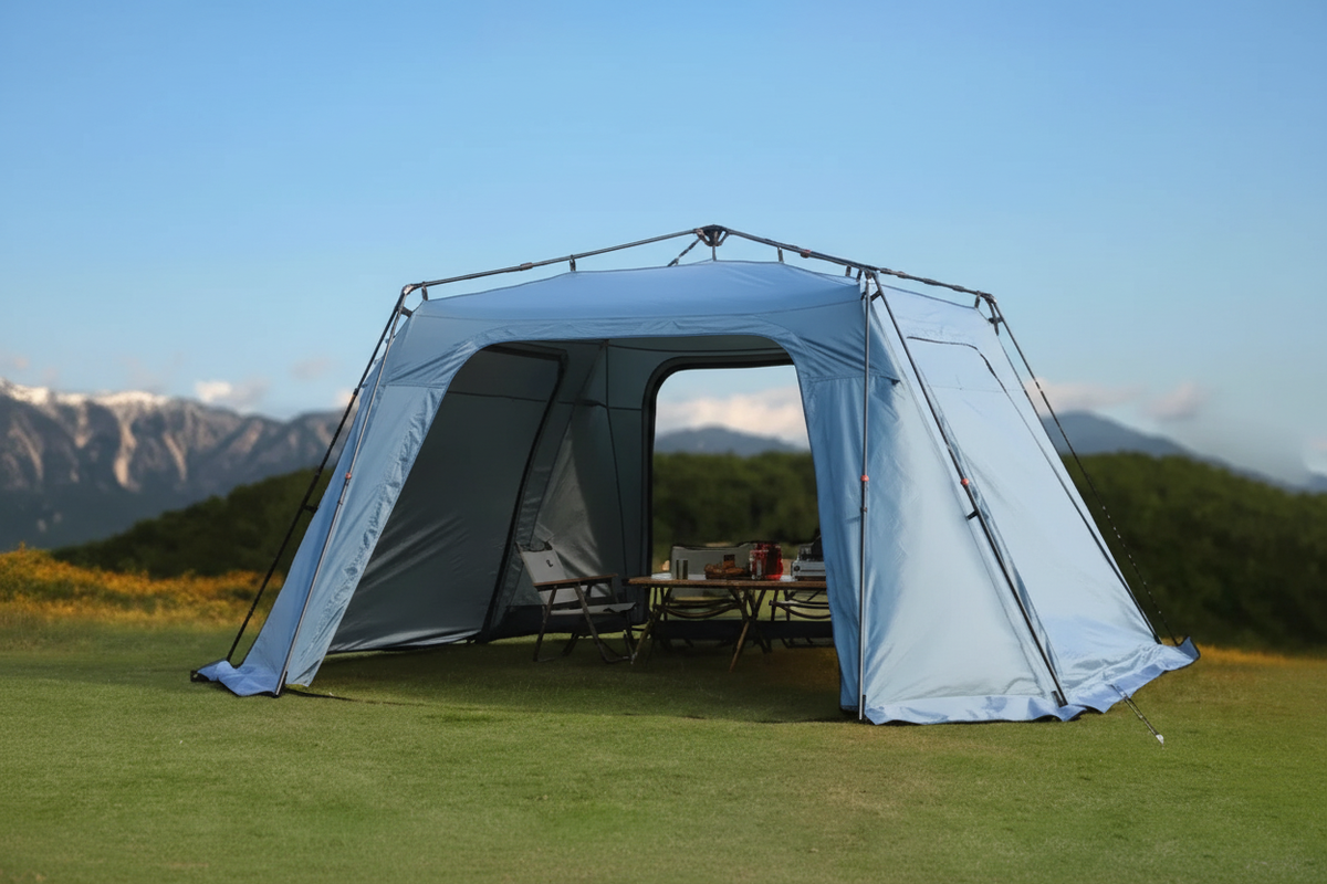 Light blue automatic dome canopy tent on green grass with dramatic mountain range backdrop, camping table and chairs inside, no text